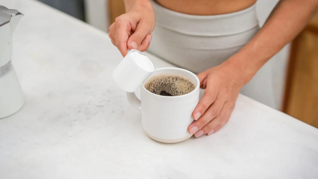 Person pouring a scoop of protein powder into a mug of coffee