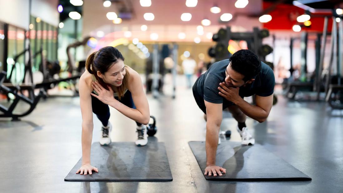 Two people look at each other while in a push-up position on exercise mats, crossing their left arms to their right shoulders